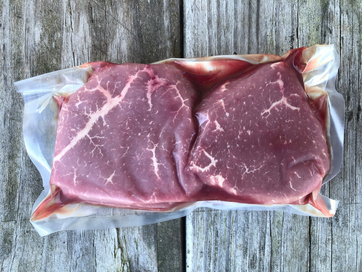 The backside of a vacuum-sealed package of Robie Farm beef eye round steaks, placed on a weathered gray wooden surface.