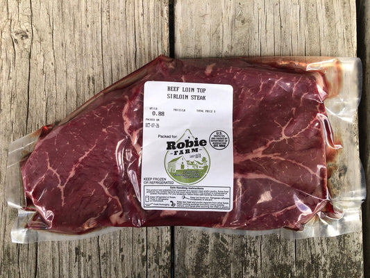 A vacuum-sealed package of Robie Farm Beef Loin Top Sirloin Steak placed on a rustic wooden surface. The package has a white label with the Robie Farm logo and product details.