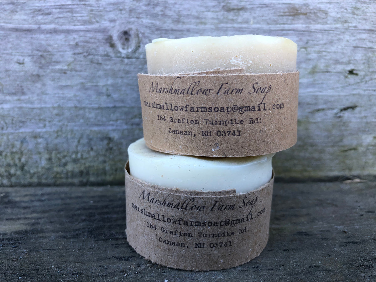 Two round Marshmallow Farm shaving pucks 
 stacked on a rustic weathered wood background, each wrapped in a brown kraft paper band with the brand name and contact details printed on it.