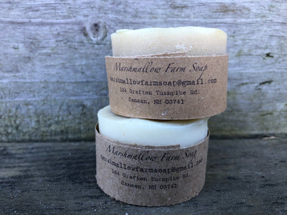 Two round Marshmallow Farm shaving pucks 
 stacked on a rustic weathered wood background, each wrapped in a brown kraft paper band with the brand name and contact details printed on it.