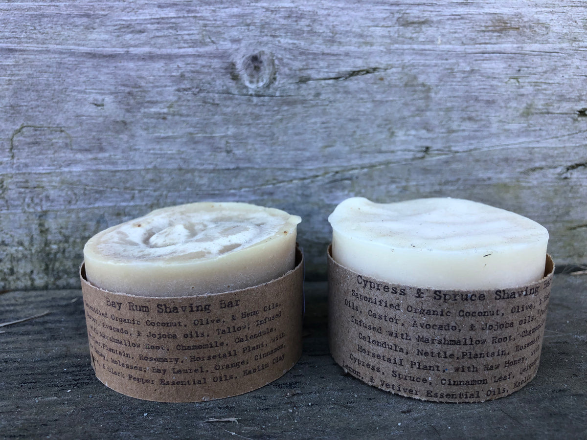 Two handmade shaving bars in brown paper bands with printed text, set against a rustic gray wooden background.