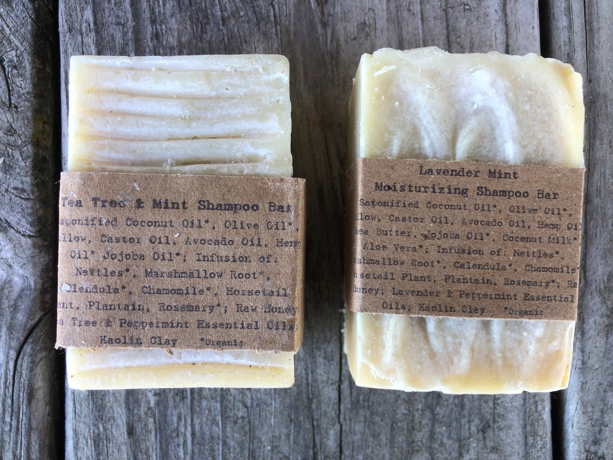 Two handmade shampoo bars wrapped in brown paper labels, displayed side-by-side on a dark rustic wooden background. The left bar is labeled 'Tea Tree & Mint Shampoo Bar' and the right bar is labeled 'Lavender Mint Moisturizing Shampoo Bar'.