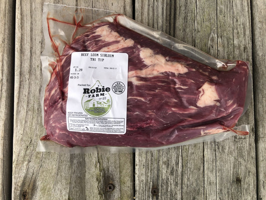 A vacuum-sealed beef loin sirloin tri tip steak with a white Robie Farm label, placed on a weathered gray wooden plank surface.
