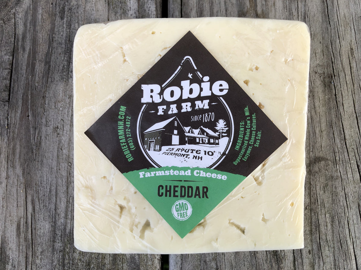 A square block of Robie Farm Farmstead  Cheddar cheese, wrapped in clear plastic with a black and green diamond-shaped label, set on a rustic gray wooden background.