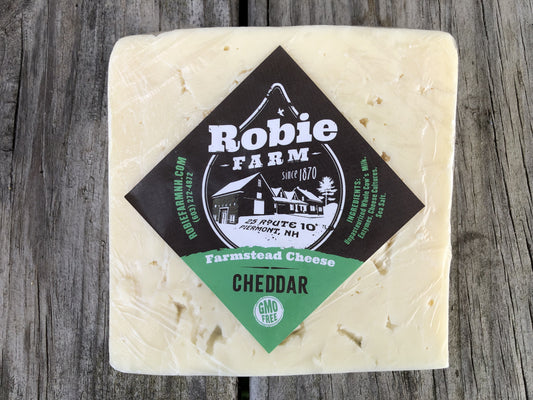 A square block of Robie Farm Farmstead  Cheddar cheese, wrapped in clear plastic with a black and green diamond-shaped label, set on a rustic gray wooden background.