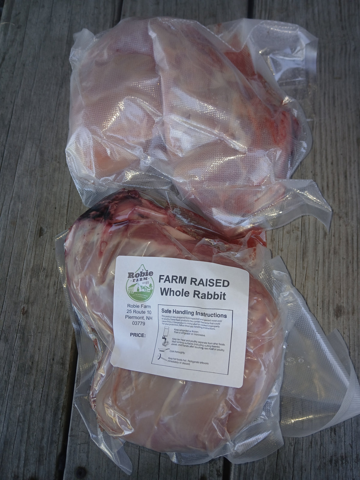 Two vacuum-sealed packages of whole rabbit meat on a weathered gray wooden surface in Piermont, NH. The lower package has a white label reading 'FARM RAISED Whole Rabbit'