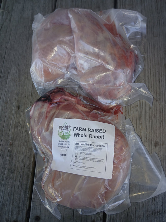 Two vacuum-sealed packages of whole rabbit meat on a weathered gray wooden surface in Piermont, NH. The lower package has a white label reading 'FARM RAISED Whole Rabbit'